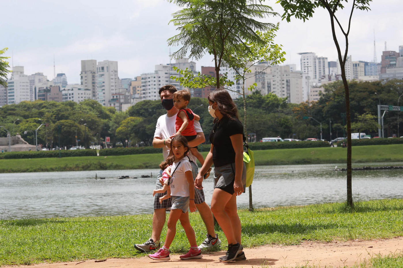 Casal de máscara caminha diante de corpo d'água cercado de grama verde; homem segura bebê nos braços e mulher segura mão de criança pequena que caminha, ambos usam máscaras